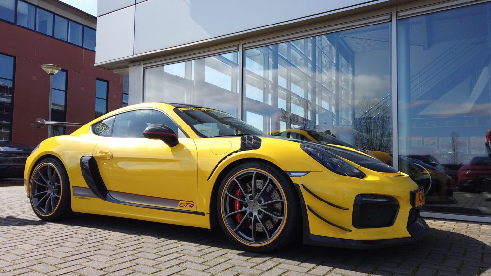 Een gele Porsche Cayman GT4 met grijze velgen en gele striping